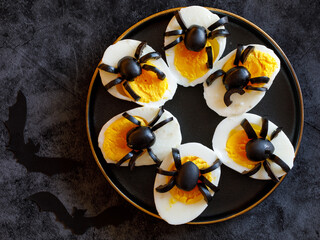 The idea for decorating a table for Halloween funny snack: stuffed eggs with spiders from olives on black plate on black background with bats, horizontal, top view flat lay