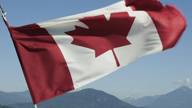 The Canadian flag waving in the wind against a blue sky with green mountains in the distance.