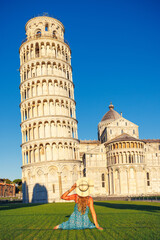 Young woman with blue dress and hat sitting on green grass looking at leaning Pisa tower- Italy