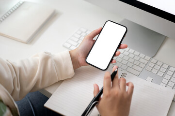 A female using her smartphone and taking notes on her notebook at her computer desk.