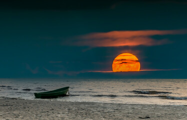 Coastal landscape during colorful rise of super-moon above cloudy horizon with anchored fishing...