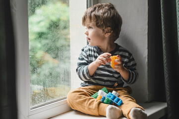 Little boy sitting on the window sill and  playing with lots of colorful plastic blocks constructor. Boy playing with construction blocks at kindergarten. 