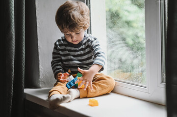 Little boy sitting on the window sill and  playing with lots of colorful plastic blocks...
