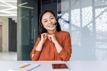 Webcam view, online video call remote meeting, businesswoman with headset phone smiling and looking at camera, asian woman talking with colleagues using laptop while sitting inside office. - Powered by Adobe