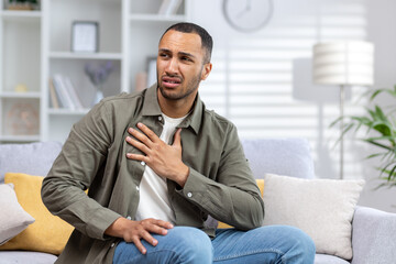 Excited young african american man sitting on sofa at home and holding chest, feeling severe pain...