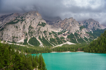 Braies lake surrounded by pine forests and the rocky ranges of the Dolomites in cloudy day, Italy.