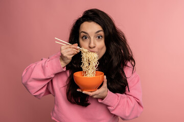 Waist-up portrait of a lady slurping down noodles out of the ceramic bowl using a pair of chopsticks while looking at the camera - obrazy, fototapety, plakaty