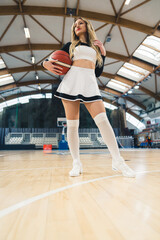 Fototapeta na wymiar Low angle shot of young woman in a black and white cheerleader uniform standing on court indoor with a basketball . High quality photo