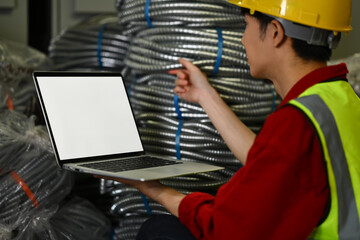 Side view of Asian male worker wearing hardhats using laptop checking stock and order details in warehouse