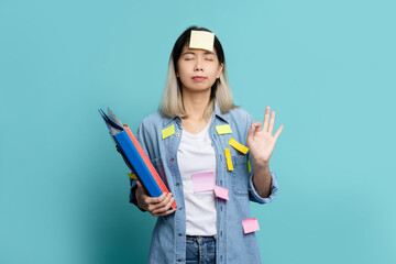Asian female office worker wearing casual clothes holding document file and hands in yoga om aum gesture meditate isolated on pastel plain light blue background.