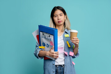 Tired and unhappy asian woman holding many document file has deadline with cup of coffee on blue...