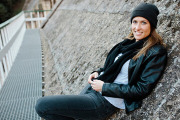 Smiling woman sitting on wall of dam. Beautiful young female wears dark winter clothes and looks at camera. Wears coat, scarf and wool hat. Copy space. Lifestyle concept.