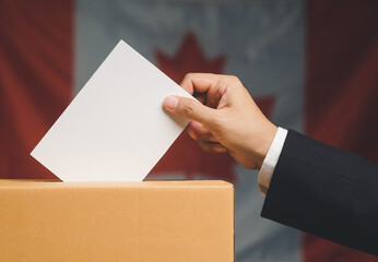 Hand voter holding ballot paper putting into the voting box at place election against the Canada flag background