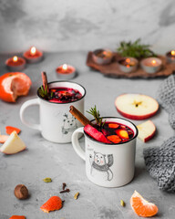 Two light cups with mulled wine, fruits, spices, candles, a piece of knitted fabric around. Christmas themed alcoholic drinks with New Year attributes.