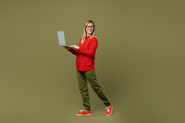 Full body side profile view young IT woman she wears red shirt casual clothes glasses hold use work on laptop pc computer look aside isolated on plain pastel green background studio. Lifestyle concept