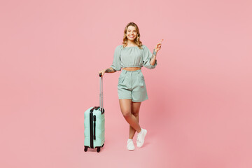 Traveler woman wears casual clothes hold suitcase point aside isolated on plain pastel pink background studio. Tourist travel abroad in free spare time rest getaway. Air flight trip journey concept.