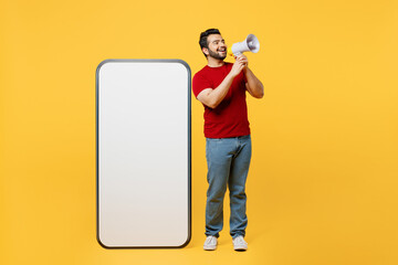 Full body young Indian man in red t-shirt casual clothes big huge blank screen mobile cell phone smartphone with area scream in megaphone isolated on plain yellow orange background Lifestyle concept