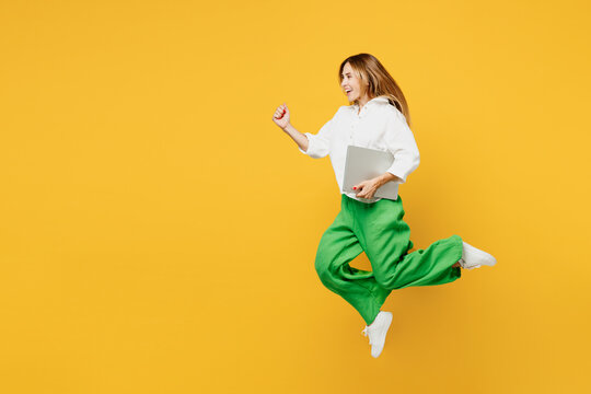 Full body young caucasian happy fun IT woman wears white shirt casual clothes jump high hold closed laptop pc computer run fast isolated on plain yellow background studio portrait. Lifestyle concept.