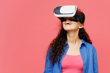 Side view young happy smiling woman of African American ethnicity she wear blue shirt casual clothes watching in vr headset pc gadget isolated on plain pastel pink background studio Lifestyle concept