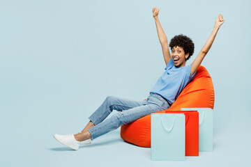 Young fun woman wears casual clothes raise up hands near paper package bags after shopping sit in bag chair do winner gesture isolated on plain blue cyan background. Black Friday sale buy day concept