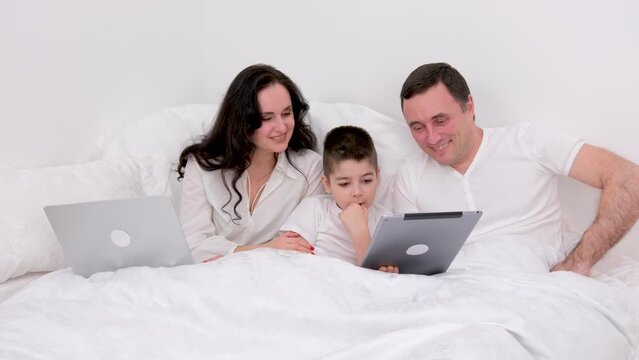 husband wife in bed with little son looking at tablet cartoon interesting game pictures laughing talking communication real people at home love life happy family life joyful people white background