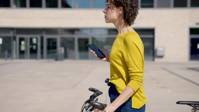 Young woman with smartphone pushing bike on Campus