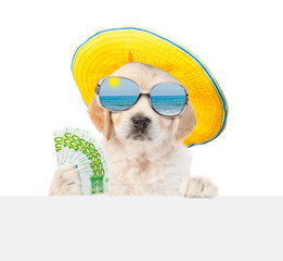 Funny Golden retriever puppy wearing mirrored sunglasses and summer hat holds Euro in it paw and looking above empty white banner. Isolated on white background