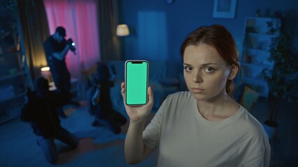 Shot of two burglars, intruders, being caught red-handed in the house. Policeman is pointing a gun at them. Woman on the foreground holding a smartphone with a mock-up, green screen.