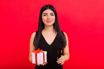 Portrait of a happy young woman with Christmas gifts and a glass of champagne on a red background.