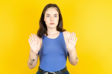 Serious Beautiful woman wearing blue tank top pulls palms towards camera, makes stop gesture, asks to control your emotions and not be nervous - obrazy, fototapety, plakaty