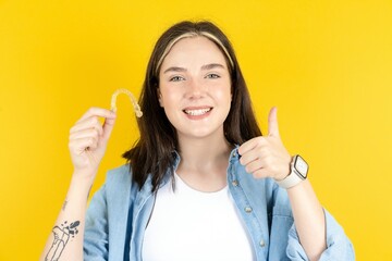 Beautiful woman wearing casual clothes  holding an invisible braces aligner and rising thumb up,...