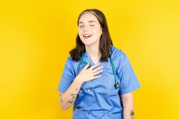 Joyful Young caucasian doctor woman wearing blue medical uniform expresses positive emotions recalls something funny keeps hand on chest and giggles happily.
