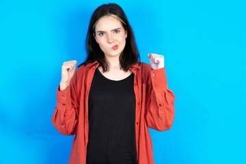 Irritated Beautiful woman wearing red jacket blows cheeks with anger and raises clenched fists...