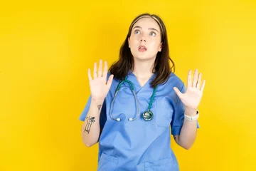 Fotobehang Young caucasian doctor woman wearing blue medical uniform keeps palms forward and looks with fright above on ceiling tries to defense herself from invisible danger opens mouth. © Roquillo