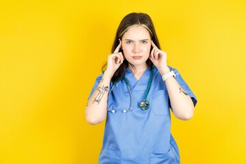 Serious concentrated Young caucasian doctor woman wearing blue medical uniform keeps fingers on temples, tries to ease tension, gather with thoughts and remember important information for exam - obrazy, fototapety, plakaty