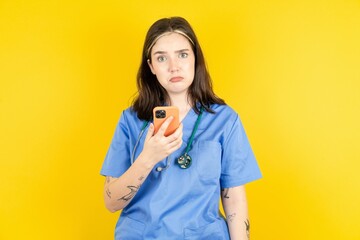 Upset dissatisfied Young caucasian doctor woman wearing blue medical uniform uses mobile software application and surfs information in internet, holds modern mobile hand