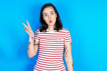 beautiful young woman wearing striped T-shirt makes peace gesture keeps lips folded shows v sign....