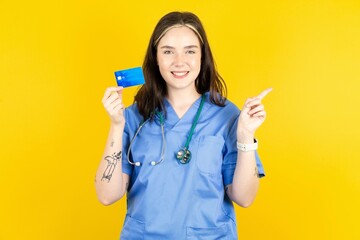 Smiling Young caucasian doctor woman wearing blue medical uniform showing debit card pointing finger empty space
