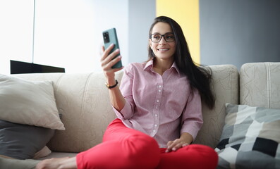 Fototapeta na wymiar Woman with glasses sits on couch smiling and looks at smartphone screen. Remote work and freelance concept