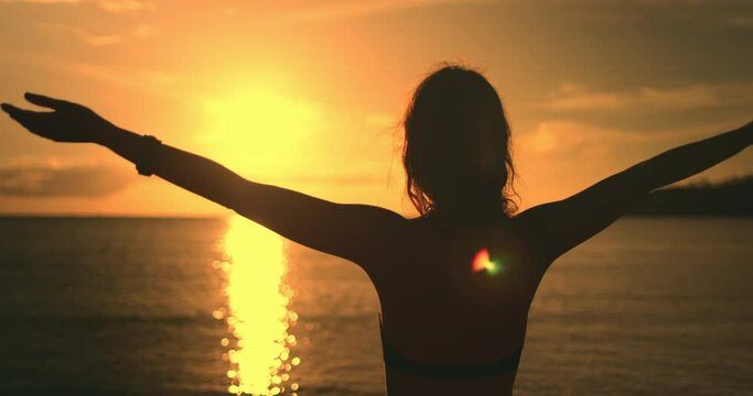Close up woman face against bright orange sunset. Girl silhouette enjoys amazing colorful sunset, standing on a tropical beach, raises hands against orange dramatic sky and ocean waves. Holiday travel