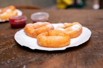 Sweet donuts fried with powder sugar on paper plate and jam in plastic bowl on wooden table. street fast food