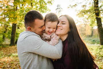 Fototapeta premium Mom, dad and daughter stand and hug on background of yellow leaves in forest. Upper half. Closeup. Young family spending time together at sunset. Autumn family photo. Happy family walks in autumn park