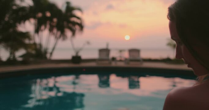 Girl silhouette near swimming pool at sunset time. Colorful sunrise sky reflected in calm water. Relax, retreat, meditation. Travel, tourism, holiday spa vacation. Slow motion portrait zooms in