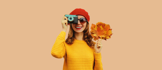 Autumn color style outfit, stylish young woman photographer with film camera holds yellow maple leaves wearing knitted sweater and red french beret hat on beige studio background