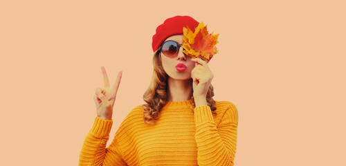 Autumn color style outfit, portrait of stylish beautiful woman model with yellow maple leaves blowing her lips sends sweet kiss wearing red french beret hat, round sunglasses on brown background