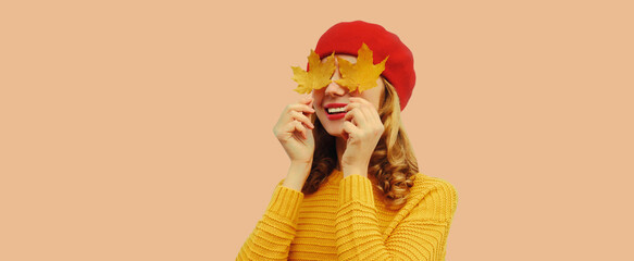 Autumn color style outfit, portrait of beautiful smiling young woman covering her eyes with yellow maple leaves wearing red french beret hat, knitted sweater on brown studio background