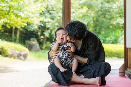 夏の緑が多い日本家屋の縁側で浴衣姿のパパに抱っこされて笑顔になる甚平姿の生後9か月の赤ちゃん