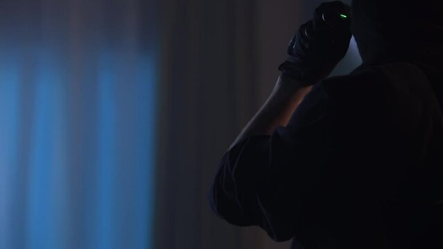 Video of a burglar going around the house with a flashlight, looking for valuables to steal. The man takes his mask off and reveals his face. Night time, blurred foreground.