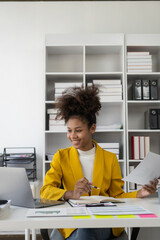 Busy professional African American female company manager wearing a business suit in a corporate office working with a laptop computer and financial accounting documents. Vertical view
