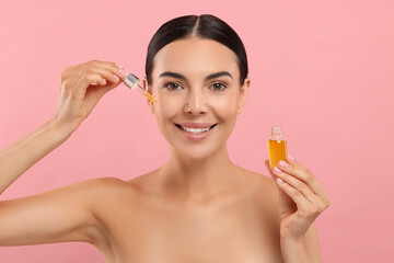 Beautiful young woman applying serum onto her face on pink background